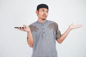 Confused young Asian Muslim man holding on smartphone with dislike expression wearing muslim clothes isolated on white background photo