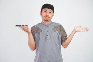 Confused young Asian Muslim man holding on smartphone with dislike expression wearing muslim clothes isolated on white background photo