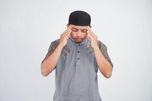 joven asiático musulmán hombre participación su cabeza sufrimiento dolor de cabeza vistiendo gris musulmán ropa aislado en blanco antecedentes foto