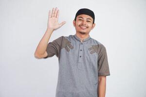 smiling or happy young asian muslim man open hand while waving hand say hello wearing grey muslim clothes isolated on white background photo