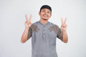 smiling or happy young asian muslim man showing pointing up with fingers number two while confident and showing peace sign with fingers wearing koko clothes isolated on white background photo