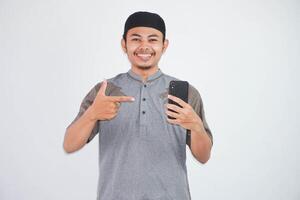 smiling asian muslim man holding and finger pointing mobile phone wearing grey muslim clothes isolated on white background photo