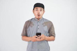 shocked young asian muslim man holding mobile phone wearing muslim clothes isolated on white background photo