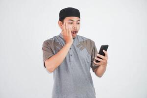 shocked young Asian Muslim man using a mobile phone, received good news wearing grey muslim clothes isolated on white background photo