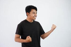 Confident cheerful handsome asian man lifting hands up wearing black t shirt winner gesture clenching fists. feels happiness show fist up success isolated on white background photo