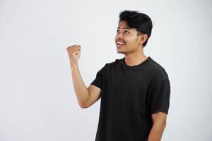 Confident cheerful handsome asian man lifting hands up wearing black t shirt winner gesture clenching fists. feels happiness show fist up success isolated on white background photo