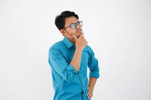 joven hermoso asiático hombre con lentes pensando y mirando para ideas mientras participación el barbilla vistiendo azul camisa aislado en blanco antecedentes foto