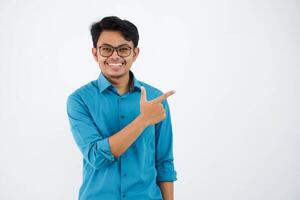 hermoso asiático masculino empleado con lentes sonriente en un actitud dedo señalando a el lado vistiendo azul camisa aislado en blanco antecedentes foto