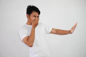 portrait of disgusted young asian man pinches nose with fingers hands looks with disgust something stinks bad smell situation wearing white t shirt isolated on white background photo