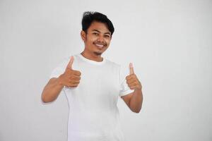 retrato de alegre asiático hombre en vistiendo blanco t camisa sonriente y demostración pulgares arriba a cámara aislado terminado blanco antecedentes foto