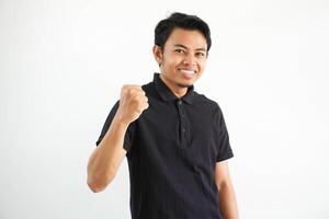 smiling or happy young asian man clenched his fists cheering carefree and excited. Victory concept, wearing black polo t shirt isolated on white background photo