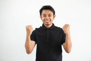 sonriente o contento joven asiático hombre apretado su puños aplausos despreocupado y entusiasmado. victoria concepto, vistiendo negro polo t camisa aislado en blanco antecedentes foto