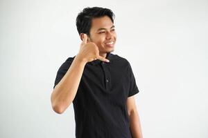 smiling young asian man posing on a white backdrop showing a mobile phone call gesture with fingers, wearing black polo t shirt. photo