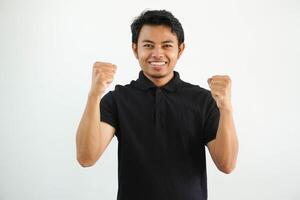 smiling or happy young asian man clenched his fists cheering carefree and excited. Victory concept, wearing black polo t shirt isolated on white background photo