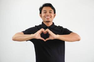 joven asiático hombre posando en un blanco fondo sonriente y demostración un corazón forma con manos, vistiendo negro polo t camisa. foto