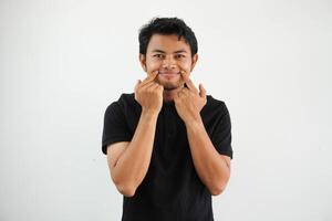 young asian man in black polo t shirt, white studio backdrop doubting between two options. force a smile with a finger. photo