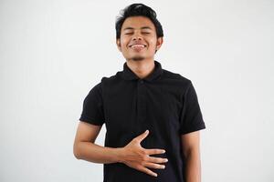 joven asiático hombre posando en un blanco fondo vistiendo negro polo t camisa toques barriguita, sonrisas suavemente, comiendo y satisfacción concepto. foto
