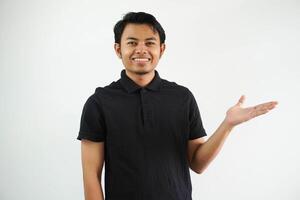 smiling young asian man in black polo shirt, white backdrop showing a copy space on a palm and holding another hand on waist. photo