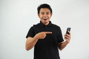 excited young asian man smiling happy while pointing to mobile phone that he hold wearing black polo t shirt isolated on white background photo