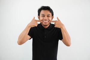 smiling young asian man pointing to his mouth with happy expression wearing black polo t shirt isolated on white background photo