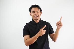 emocionado joven asiático hombre sonriente confidente con ambos mano señalando a el izquierda lado vistiendo negro polo t camisa aislado blanco antecedentes foto