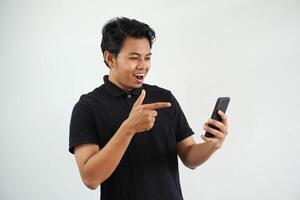 excited young asian man smiling happy while pointing to mobile phone that he hold wearing black polo t shirt isolated on white background photo