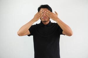 joven asiático hombre posando en un blanco fondo temeroso cubierta ojos con manos, vistiendo negro polo t camisa. foto