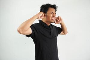 enojado joven asiático hombre cerca su orejas con dedos vistiendo negro polo t camisa aislado en blanco antecedentes foto