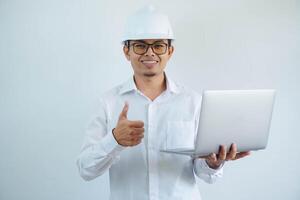 Young asian architect man wearing builder safety helmet over isolated background doing happy thumbs up gesture and holding laptop with hand. photo