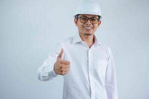 Young asian architect man wearing builder safety helmet over isolated background doing happy thumbs up gesture with hand. Approving expression looking at the camera showing success. photo