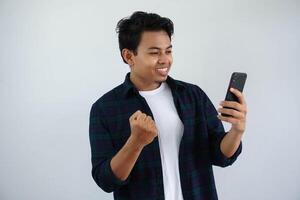 young asian man clenching fist showing excitement when looking to his phone isolated on white background photo