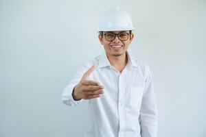 arquitecto joven asiático hombre vistiendo blanco casco en pie terminado aislado blanco antecedentes sonriente simpático ofrecimiento apretón de manos como saludo y acogedor exitoso negocio. foto