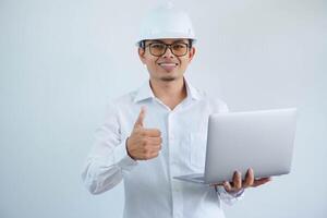 Young asian architect man wearing builder safety helmet over isolated background doing happy thumbs up gesture and holding laptop with hand. photo