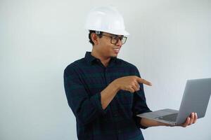 smiling young asian architect man wearing builder safety helmet pointing his laptop isolated on white background photo