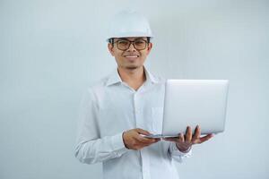 Young asian architect man wearing builder safety helmet over isolated background doing happy with holding laptop with hand. photo