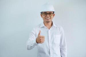 Young asian architect man wearing builder safety helmet over isolated background doing happy thumbs up gesture with hand. Approving expression looking at the camera showing success. photo