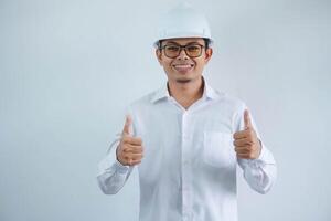 Young asian architect man wearing builder safety helmet over isolated background doing happy thumbs up gesture with hand. Approving expression looking at the camera showing success. photo