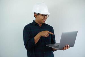 sonriente joven asiático arquitecto hombre vistiendo constructor la seguridad casco señalando su ordenador portátil aislado en blanco antecedentes foto