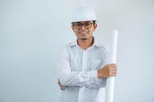 joven asiático hombre es ingeniero vistiendo casco en pie con participación proyecto papel plan y sonriente con seguro, arquitecto o contratista, trabajador o mano de obra, industrial concepto. foto