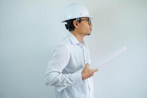 side view smiling young Asian architect wearing helmet hardhat and holding project paper plan isolated on white background. photo