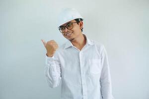 Young asian architect man wearing builder safety helmet over isolated background smiling with happy face looking and pointing to the side with thumb up. photo