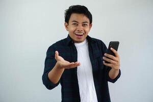 Young Asian man looking to camera showing wow expression while holding handphone isolated on white background photo