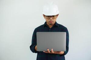 grave joven asiático arquitecto hombre vistiendo constructor la seguridad casco mecanografía en el ordenador portátil aislado en blanco antecedentes foto