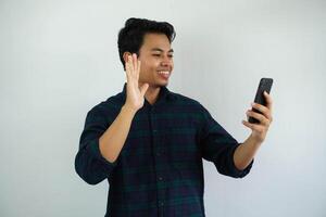 sonriente joven asiático hombre ondulación mano con contento cara cuando haciendo vídeo llamada con alguien aislado en blanco antecedentes. foto