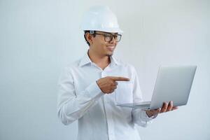 smiling young asian architect man wearing builder safety helmet pointing his laptop isolated on white background photo