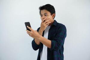 joven asiático hombre demostración conmocionado facial expresión con mano cubierta boca mientras mirando a su móvil teléfono aislado en blanco antecedentes foto