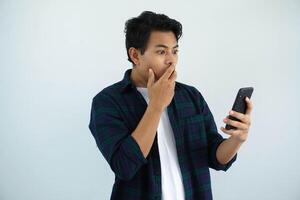 joven asiático hombre demostración conmocionado facial expresión con mano cubierta boca mientras mirando a su móvil teléfono aislado en blanco antecedentes foto