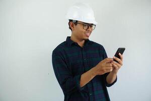 smiling young Asian male engineer wearing white hard hat holding mobile phone for construction work isolated on white background. photo