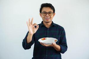 joven asiático hombre sonriente contento y dar Okay firmar con mano mientras demostración vacío cena plato aislado en blanco antecedentes. foto