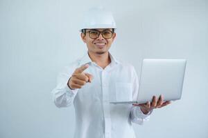 smiling young asian architect man wearing builder safety helmet holding laptop and pointing forward isolated on white background photo